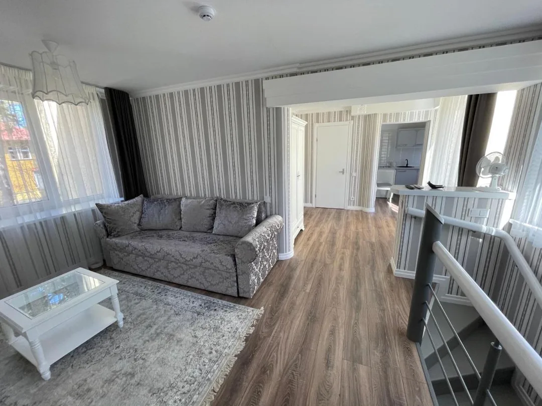 Svetainė su sofa ir kavos staliuku, gilumoje — virtuvė ir laiptų turėklai