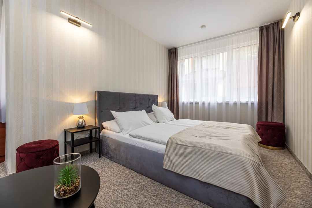 Superior double room: wide bed with a grey headboard, bedside table with a lamp, window and a round table